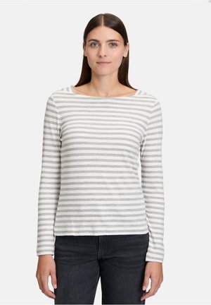 Donna con capelli castano lisci, che indossa una maglietta a maniche lunghe bianca e grigia a righe e pantaloni scuri, in piedi davanti a uno sfondo neutro.