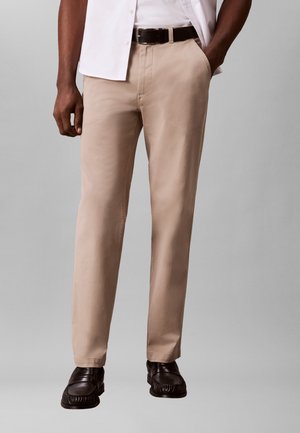 Mand iført beige bukser med lige ben, sort bælte og sorte loafers, med den ene hånd i lommen, mod en ensfarvet grå baggrund.