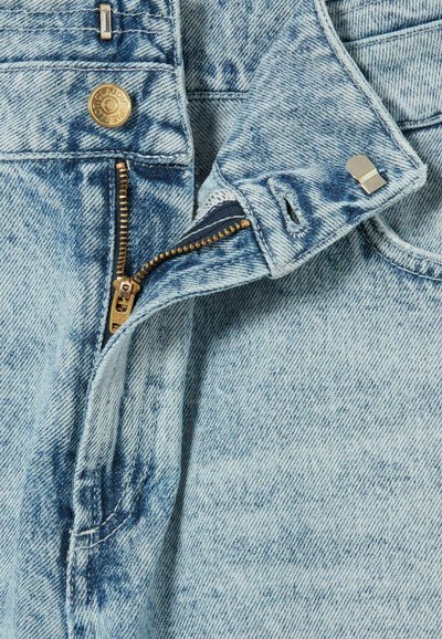 Denim blu chiaro con una texture sbiadita, caratterizzato da una cerniera in ottone, cuciture visibili e dettagli in metallo vicino alla vita.