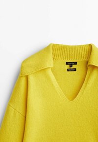 Geel wollen V-halstrui met een gestructureerde gestreepte kraag en schouderaccenten, een gladde body en labels met informatie over het materiaal en verzorgingsinstructies.