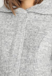 Cappotto in lana grigia, texture con cappuccio grande e cuciture visibili. Presenta una chiusura a bottone e dettagli delle cuciture curve. Materiale morbido e caldo.