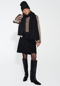 Berretto di maglia nero, sciarpa nera oversized, blouse beige lucido, gonna nera aderente, stivali neri in pelle al ginocchio con tacco a zeppa.