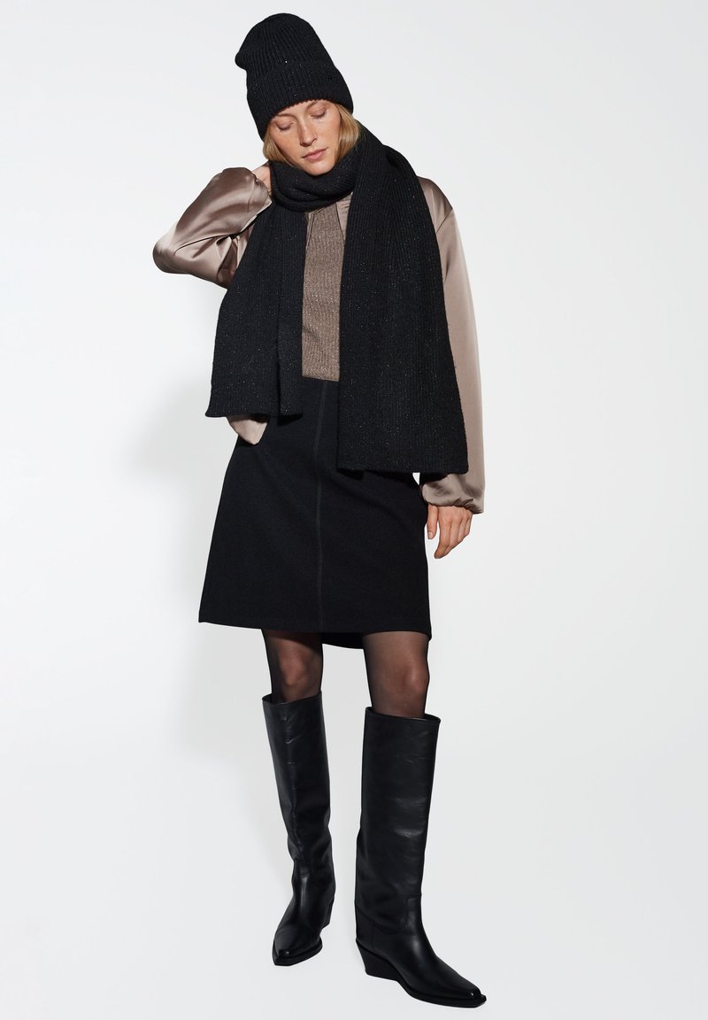 Berretto di maglia nero, sciarpa nera oversized, blouse beige lucido, gonna nera aderente, stivali neri in pelle al ginocchio con tacco a zeppa.