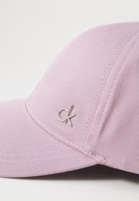 Lys rosa baseball-lue med teksturert stoff og buet skjerm. Har et sølvbrodert "ck"-logo foran.