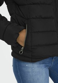 Svart puffjakke med horisontalt quiltet design, med en sidelommer med glidelås og en glatt tekstur. Brukt med blå denimjeans.