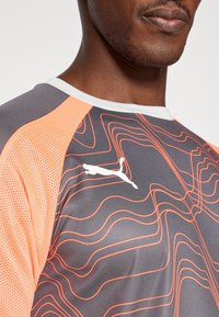 Sivá športová košeľa s oranžovými sieťovinovými akcentmi, s vlnitým vzorom v oranžovej farbe a bielym logom Puma na ľavom hornom rohu.