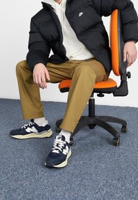 Persona con chaqueta negra de Nike, pantalones beige, calcetines blancos y zapatillas de tenis New Balance azul marino, sentado en una silla de oficina naranja sobre una alfombra azul.
