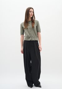 Cardigan verde khaki a maniche corte con bottoni, abbinato a pantaloni neri a vita alta plissettati e scarpe nere a punta.