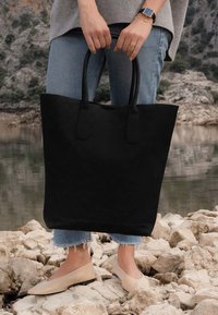 Zwarte suede schoudertas met dubbele handvatten, met een soepele textuur en minimalistisch ontwerp. Draagt door een persoon op rotsachtig terrein.