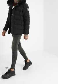 Person som går iförd en svart pufferjacka med pälsfodrad huva, maroon- och vitrandig skjorta, grå slim fit-jeans och svarta sneakers.