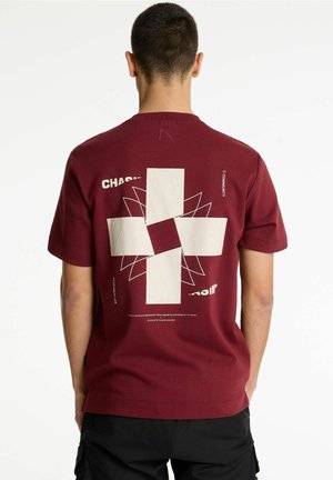 Homme portant un t-shirt bordeaux avec une grande croix blanche abstraite et du texte dispersé au dos, associé à un pantalon noir.