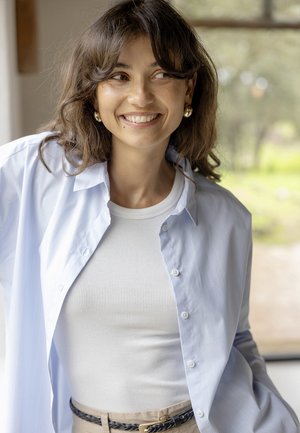 Fiatal nő középhosszú barna hajjal mosolyog, fehér bordás felsőt, világoskék gombos inget, bézs övet és arany karika fülbevalókat visel.