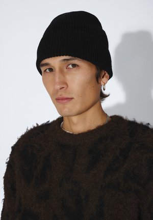 Ung mand iført en sort strikket hue, en blød mørkebrun sweater og en sølv ørering i hoop-stil mod en ensfarvet lys baggrund.