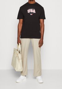 Black cotton t-shirt featuring "USA" graphic, beige plaid trousers, white sneakers, and a cream tote bag. Casual design, relaxed fit.