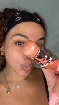 Vrouw met krullend haar en zwarte hoofdband houdt een bronskleurige make-up stick dicht bij haar gezicht en kijkt naar de camera.