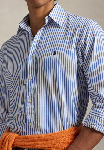 Ριγέ πουκάμισο σε μπλε και λευκό με ορατά κουμπιά και μικρό κεντημένο λογότυπο, συνδυασμένο με πορτοκαλί πλεκτό πουλόβερ δεμένο γύρω από τη μέση.