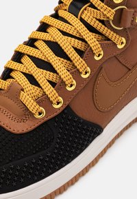 Bruine leren sneaker met zwarte accenten, gouden veterogen en gele gevlochten veters. Getextureerd zwart zijpaneel en witte rubberen zool.