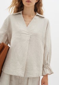 Blusa in lino, di un beige chiaro, a maniche lunghe con scollatura a V, dettagli di pieghe e polsini arricciati. La modella tiene una borsa marrone.