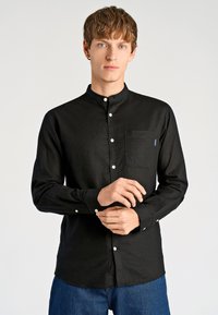 Czarna koszula z długim rękawem, kołnierzykiem typu band, białymi guzikami oraz kieszenią na piersi; wykonana z teksturowanej tkaniny, zestawiona z niebieskimi jeansami.
