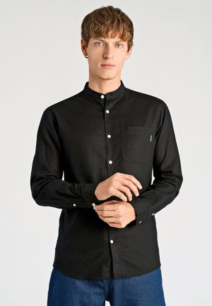 Czarna koszula z długim rękawem, kołnierzykiem typu band, białymi guzikami oraz kieszenią na piersi; wykonana z teksturowanej tkaniny, zestawiona z niebieskimi jeansami.