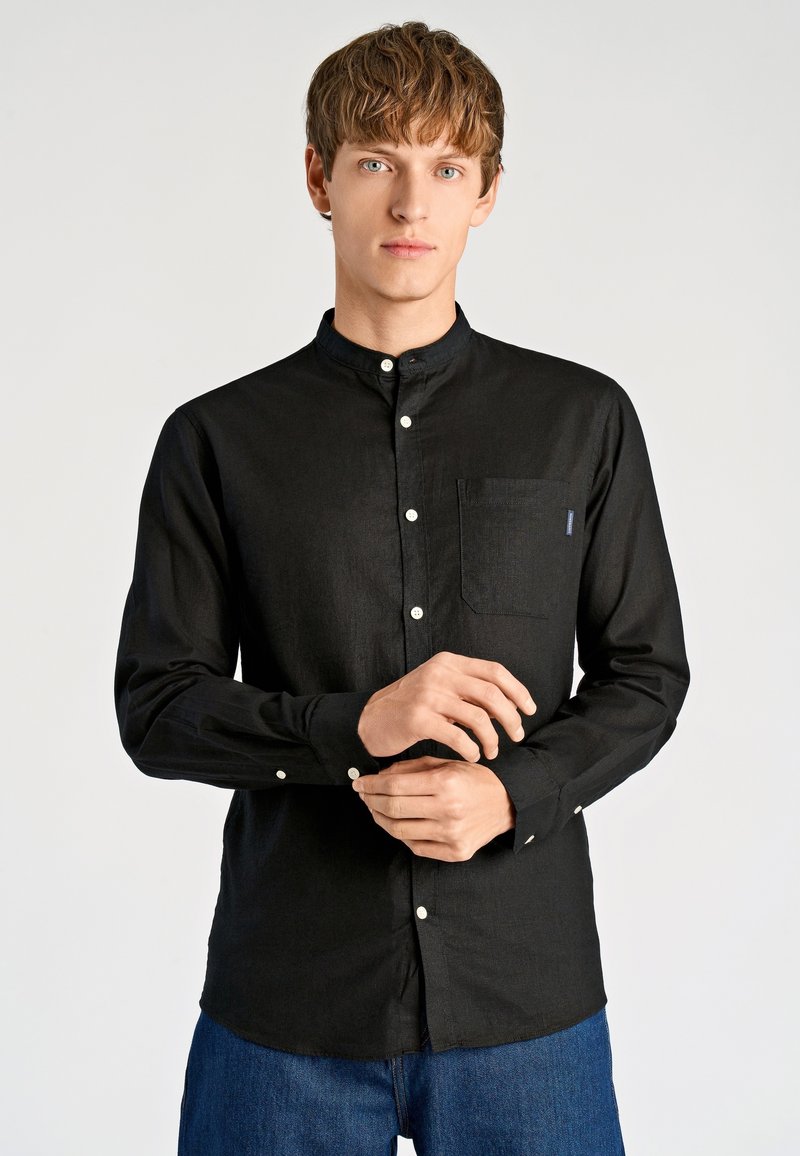 Zwart shirt met lange mouwen en een bandkraag, witte knopen en een borstzak; gemaakt van een gestructureerde stof, gecombineerd met blauwe denim jeans.