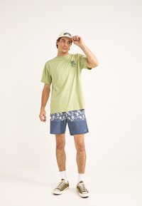 Camiseta verde claro con gráfico de palmera, combinada con pantalones cortos azul marino que presentan un patrón floral blanco. Zapatillas y gorra verde oliva.