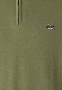 Olivno zelena polo majica s polu-zip ovratnikom, s malim vezenim logotipom krokodila u bijeloj i zelenoj boji na lijevoj strani prsnog koša. Glatka tekstura.