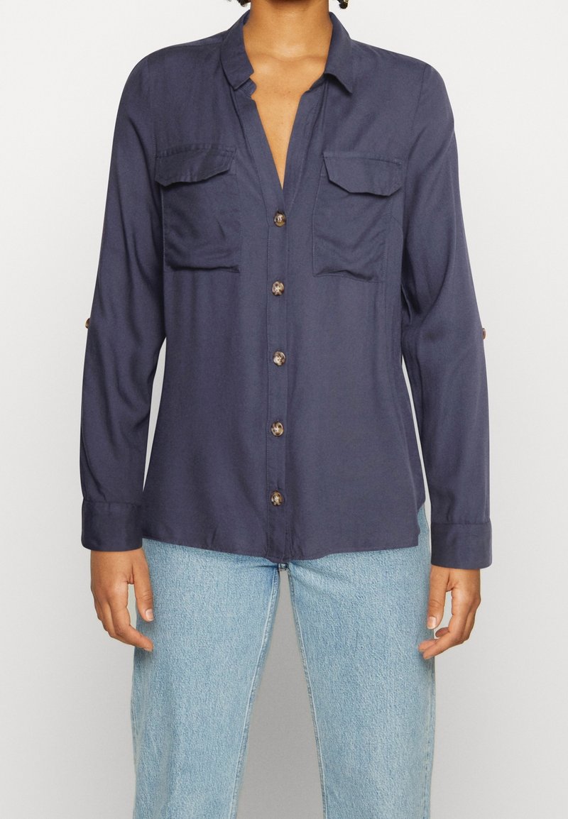 Personne portant une chemise bleu foncé à boutons avec deux poches poitrine et un jean bleu clair, montrée des épaules jusqu'aux hanches.