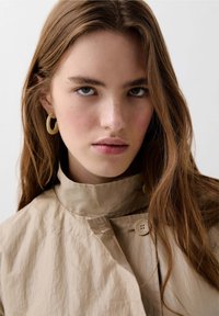 Jacket bej, ușor, cu guler înalt, detalii cu nasturi și textură moale. Modelul poartă cercei rotunzi, de culoare deschisă.