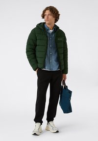 Joven vestido con una chaqueta acolchada verde oscuro, una camisa de mezclilla, pantalones negros, zapatillas blancas, sosteniendo una bolsa de tela azul, de pie contra un fondo liso.