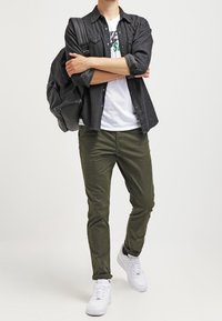 Svart skjorta med knappar över en vit t-shirt, olivgröna byxor, vita sneakers och en svart ryggsäck. Stående ställning, armarna i kors.