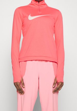 Langärmliges sportliches Oberteil in Korallenrot mit weißem Nike-Swoosh-Logo, kombiniert mit hellrosa Hose an einer Person mit dunklem Hautton.