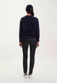 Pull en maille navy avec un motif en torsades, associé à un jean en denim foncé. La personne porte une casquette assortie et des baskets blanches.