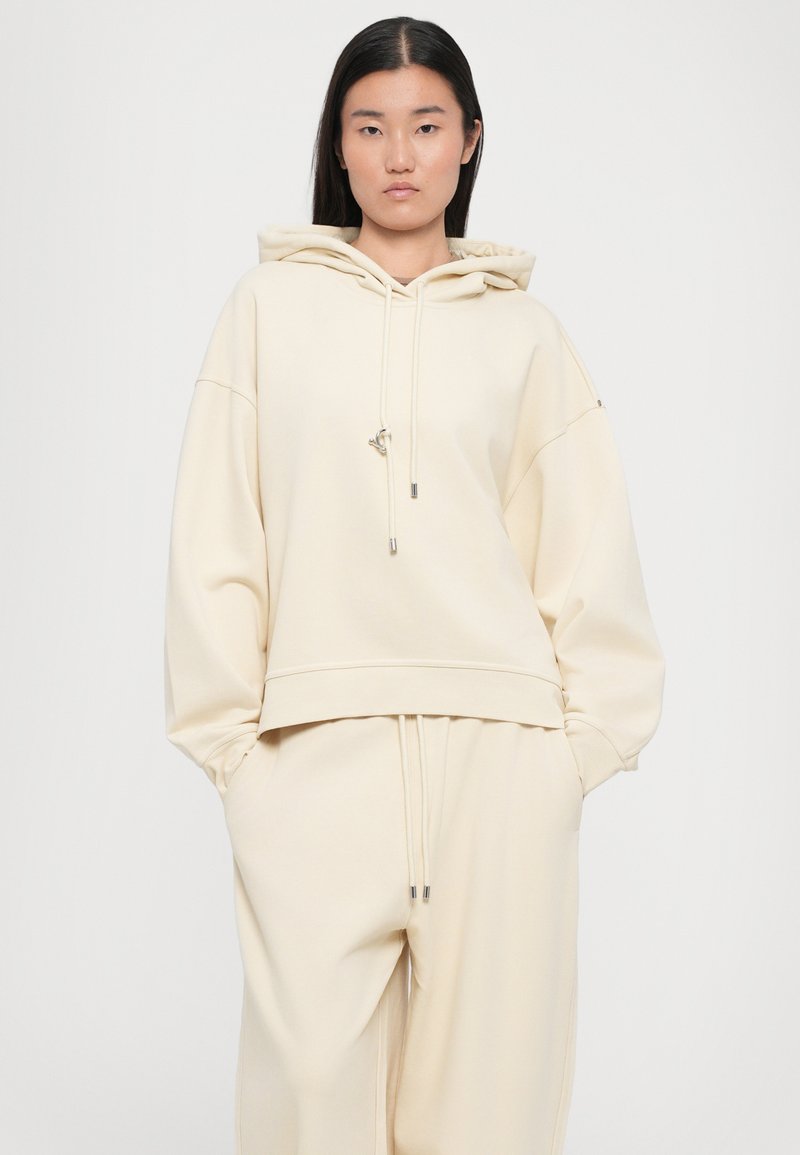 Cream-colored oversized hoodie made of soft fabric, featuring a drawstring hood and ribbed hem, paired with matching loose-fit pants.