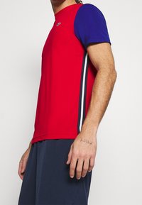 Man som bär en kortärmad tröja i röd och blå med vita och marinblå sidostreck, marinblå shorts och en liten tatuering på sin högra hand.