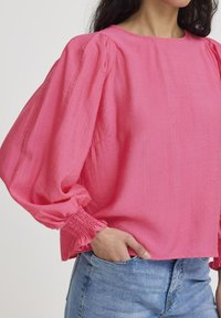 Růžová blůza s dlouhými rukávy, na ramenou nařasená a se smockovanými manžetami, vyrobená z lehké látky. S kombinací modrých džín.