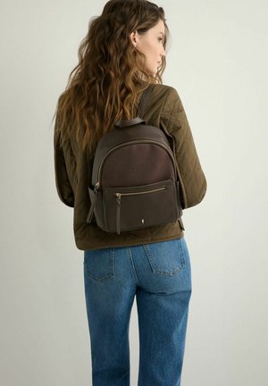 Kobieta z długimi falującymi włosami, ubrana w brązową pikowaną kurtkę i niebieskie jeansy, niosąca na plecach ciemnobrązowy skórzany plecak.