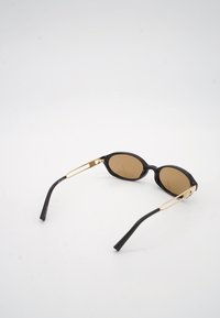 Μαύρα οβάλ γυαλιά ηλίου με καφέ φακούς και χρυσά μεταλλικά μπράτσα, τοποθετημένα σε λευκή επιφάνεια.