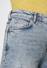 Jean en denim bleu clair avec des accessoires en cuivre, comprenant une poche avant et une ceinture texturée, porté avec un haut en tricot jaune.