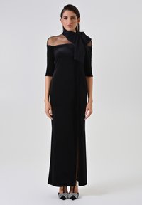 Zwarte fluwelen avondjurk met off-shoulder ontwerp, doorschijnende nekrand, driekwart mouwen, slanke pasvorm en een zijsplit, gecombineerd met versierde hakken.