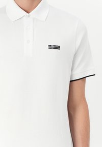 Witte poloshirt met een drieknoopsluiting, zwarte kraagaccenten en een klein geborduurd logo op de borst. Zacht katoen materiaal.