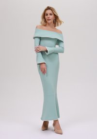 En tettsittende, off-the-shoulder mintgrønn kjole med hvit trim og klokkelignende ermer, som går ut til en flagrende underkant, kombinert med nakne hæler.