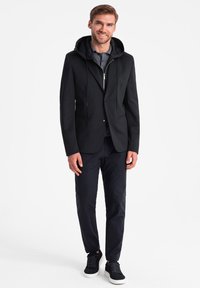 Schwarze Blazerjacke mit Kapuze, ausgestattet mit einem Reißverschluss, zwei vorderen Taschen, getragen über einem grauen Hemd und kombiniert mit dunklen Hosen und Turnschuhen.