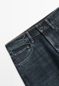 Dunkelblaue Jeans aus Denim mit Vordertasche, Gürtelschlaufen und silbernem Knopfverschluss vor weißem Hintergrund.