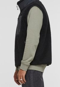 Černá fleeceová vesta s vysokým límcem, s přední kapsou na zip a texturovaným materiálem; nošena přes tlumeně zelené tričko s dlouhým rukávem.