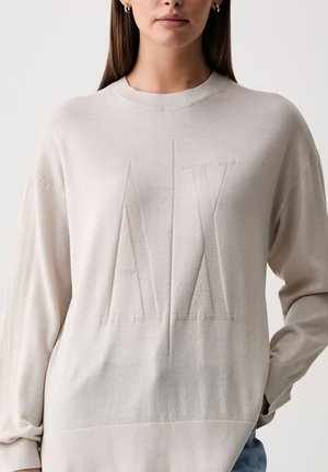 Modèle portant un sweat-shirt beige clair à col rond, orné de grandes lettres AX ton sur ton, avec un jean bleu.