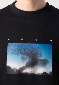 Chemise noire avec le texte "HUGO" au-dessus d'une photo d'une murmuration d'étourneaux formant des motifs tourbillonnants dans le ciel au-dessus de silhouettes d'arbres.