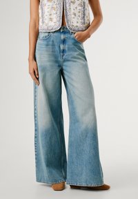 Svijetloplave široke traperice s izblijeđenim pranjem, visokog struka i klasičnim dizajnom s pet džepova. U kombinaciji s topom bez rukava s uzorkom.