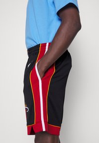 Personne portant un short de basketball noir avec des bandes rouges, blanches et jaunes et une poche latérale, associé à une chemise bleu clair.