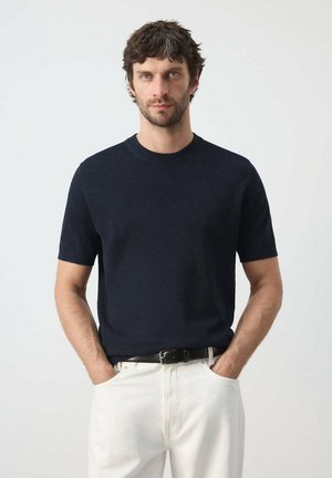 Homme aux cheveux courts, bouclés et foncés, portant un pull noir à manches courtes et un pantalon blanc, les mains dans les poches, sur un fond uni.
